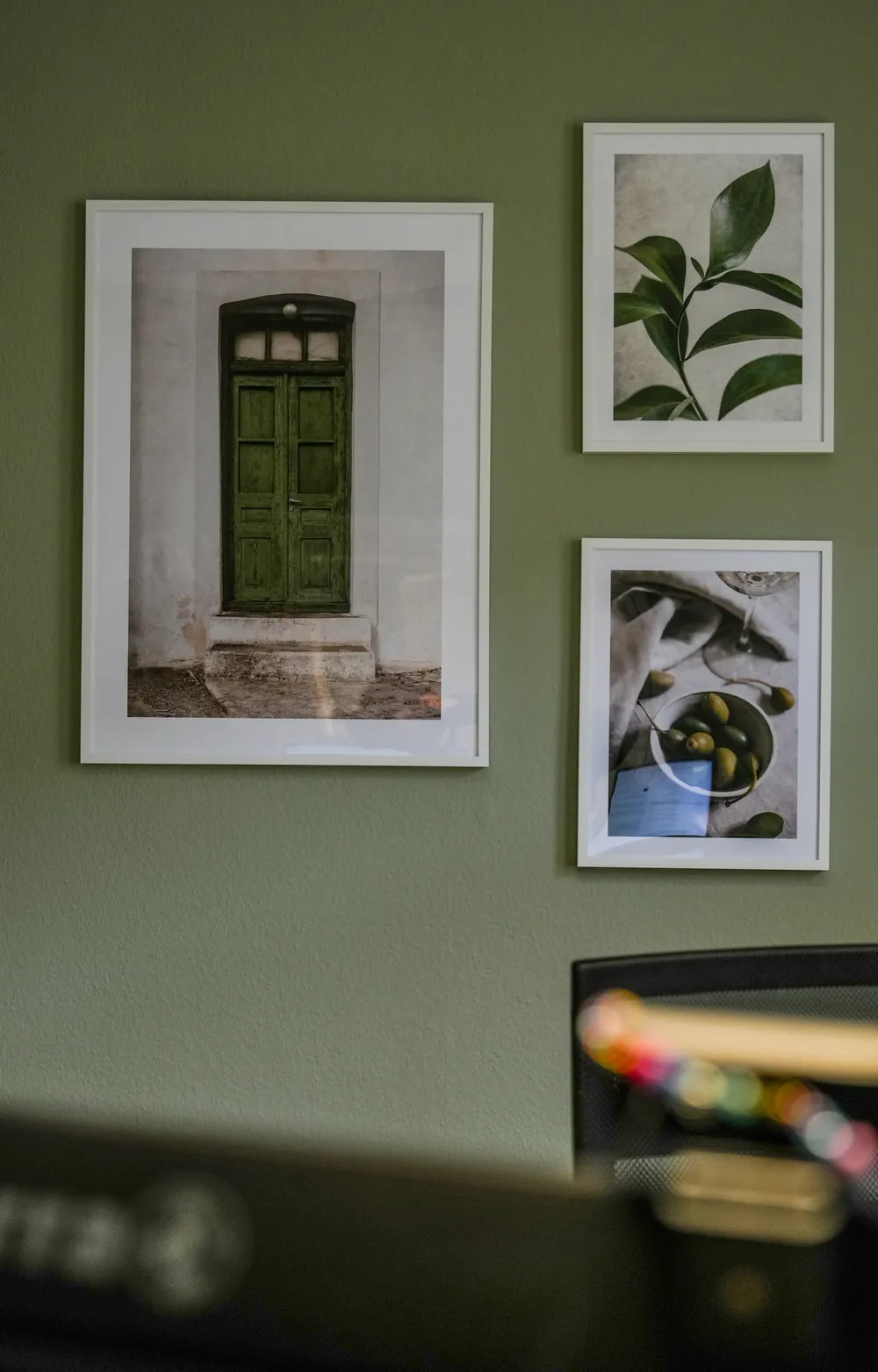 Bilderwand an olivgrüner Wand mit Fotos einer grünen Tür, eines Zweigs und einer Schale Oliven, davor ein Arbeitsplatz