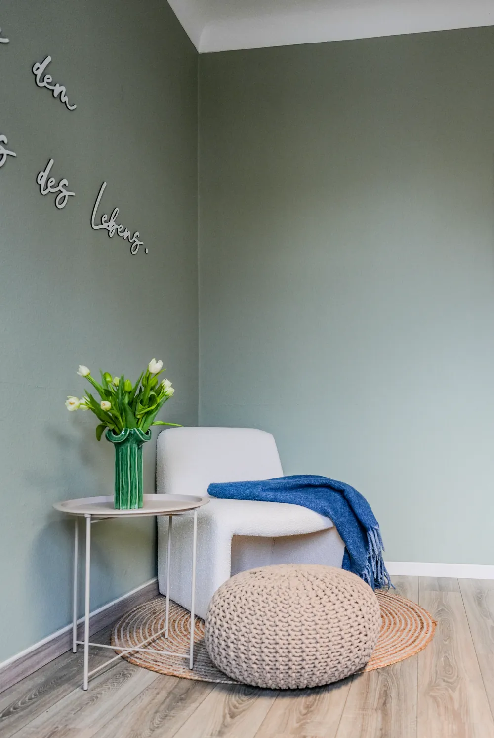 Leseecke mit weißem Sessel, blauer Decke, Strickpouf und Tulpen an salbeigrüner Wand