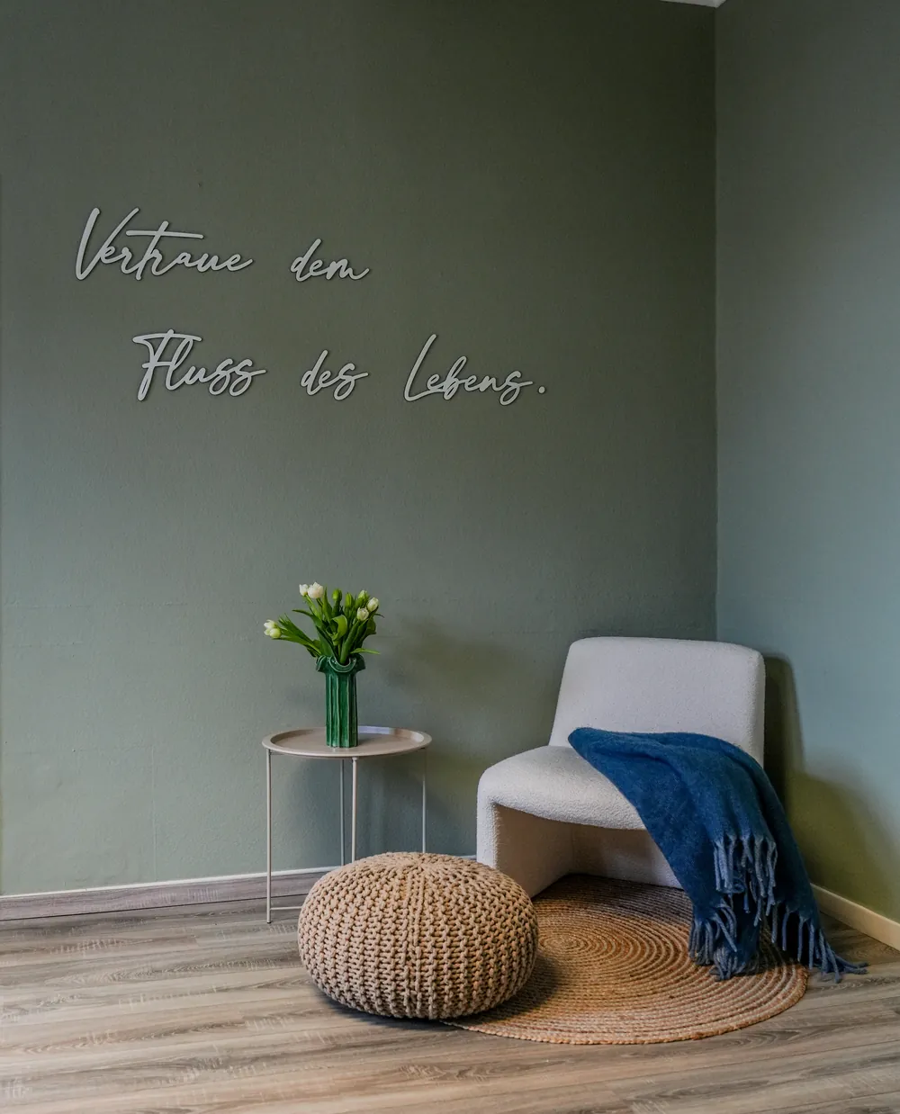 Sitzecke mit Sessel, Tulpen und dem Schriftzug Vertraue dem Fluss des Lebens an salbeigrüner Wand
